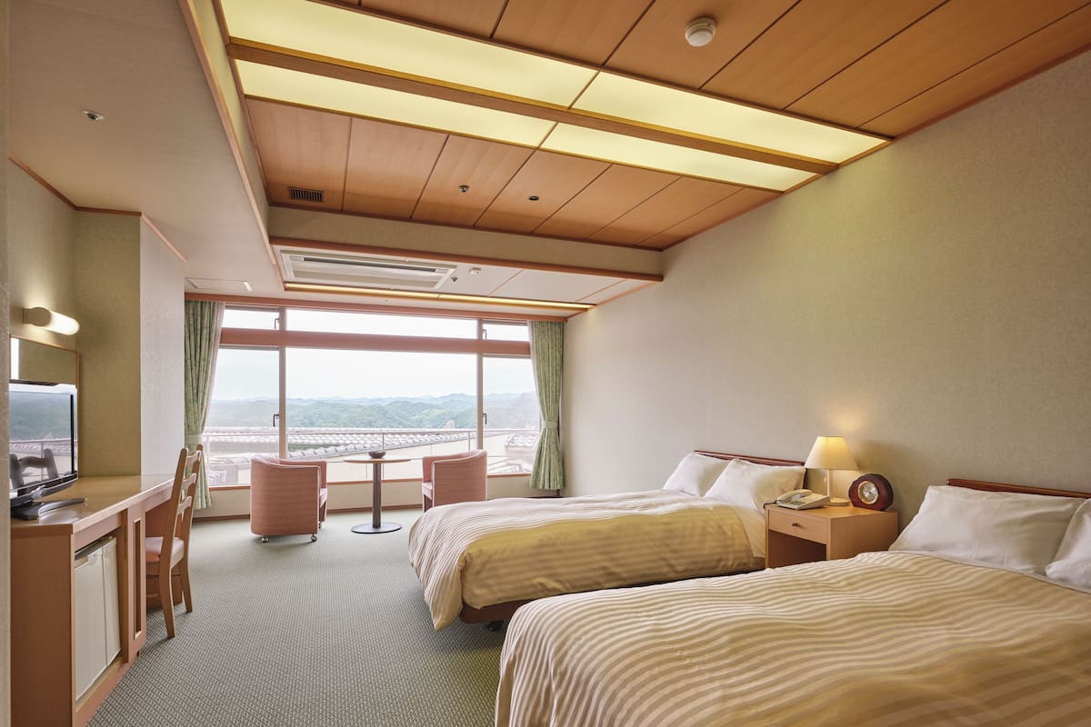 Une chambre d’hôtel avec deux lits, un bureau avec une chaise, une petite table et une grande fenêtre donnant sur les montagnes.
