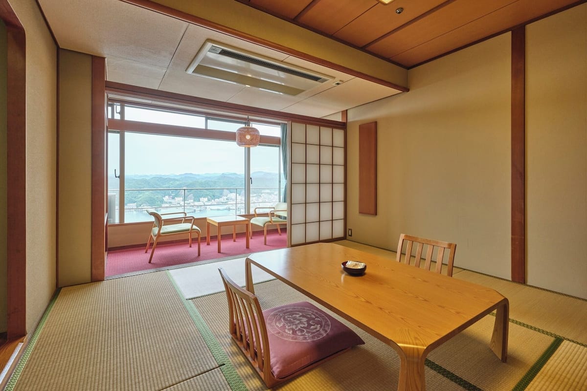 Une pièce de style japonais traditionnel, avec un sol recouvert de tatamis, une table en bois et une vue sur le paysage urbain à travers de grandes fenêtres.