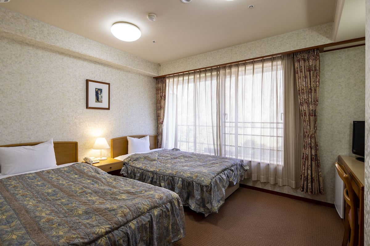 Une chambre d’hôtel avec deux lits, une table de chevet avec une lampe, une télévision et une fenêtre avec des rideaux.