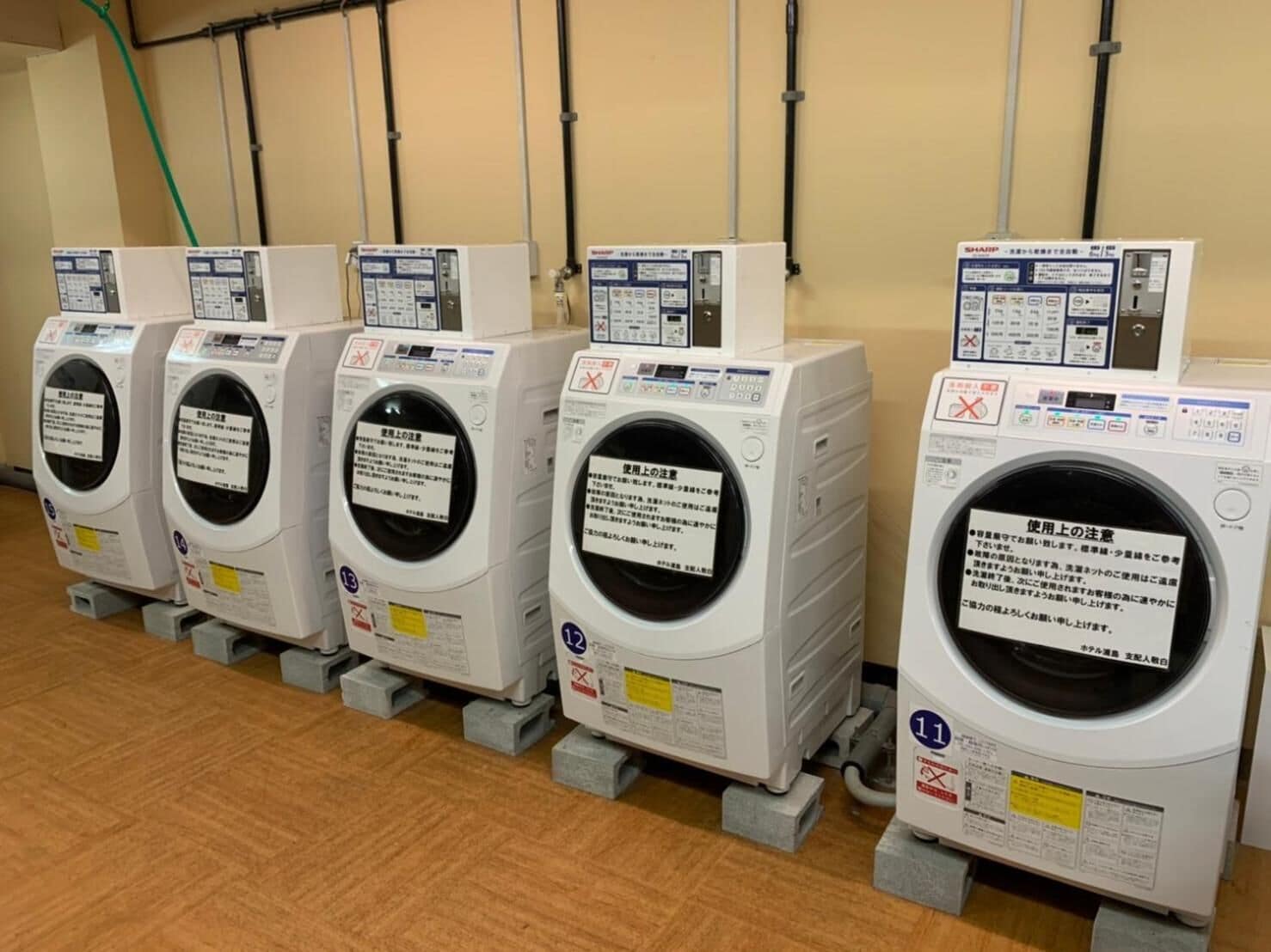 A row of white laundry machines with control panels, placed on grey platforms against a beige wall with visible pipes.