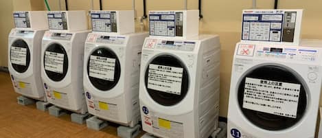 A row of white laundry machines with control panels, placed on grey platforms against a beige wall with visible pipes.