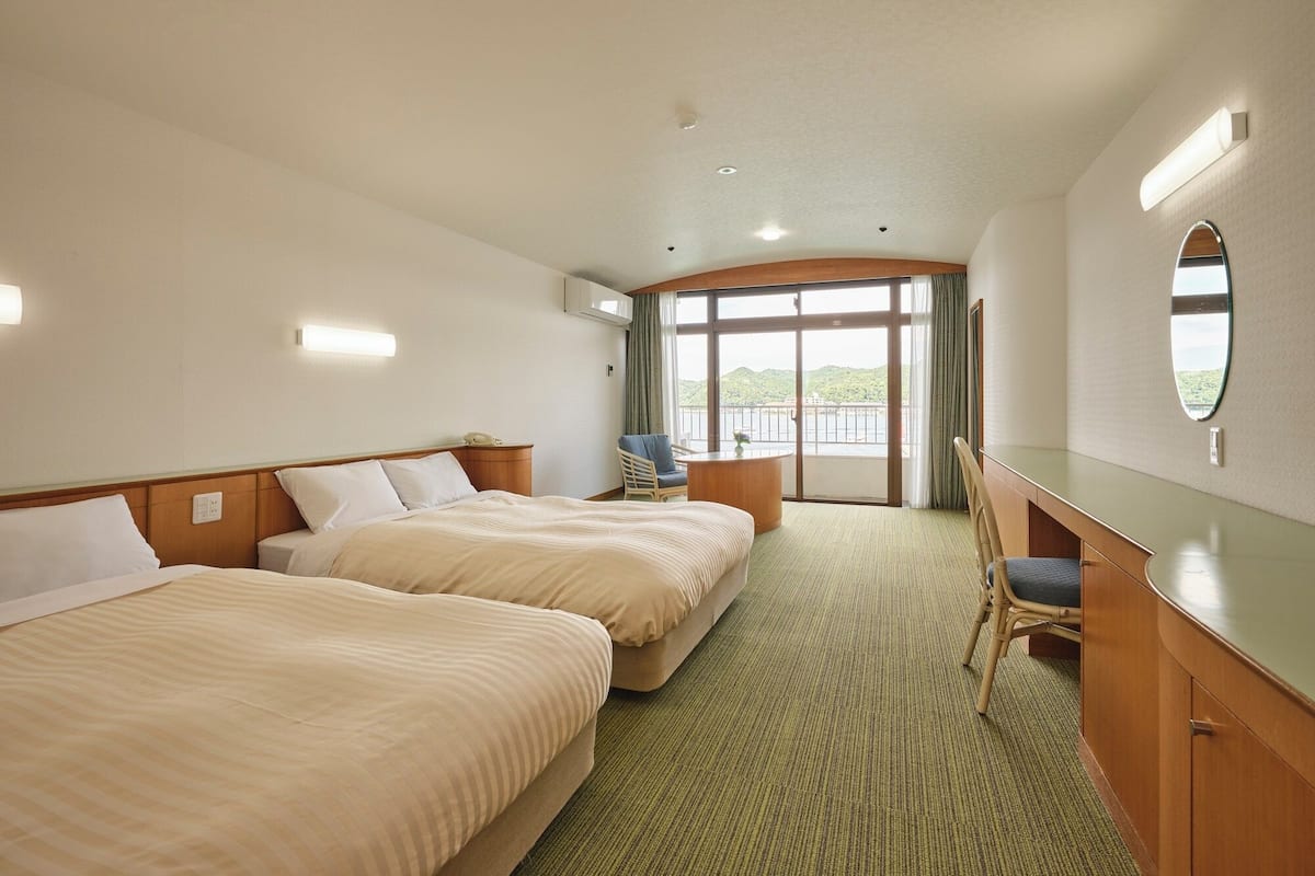 Une chambre d’hôtel avec deux lits, un bureau, une chaise et une grande fenêtre donnant sur un paysage verdoyant.