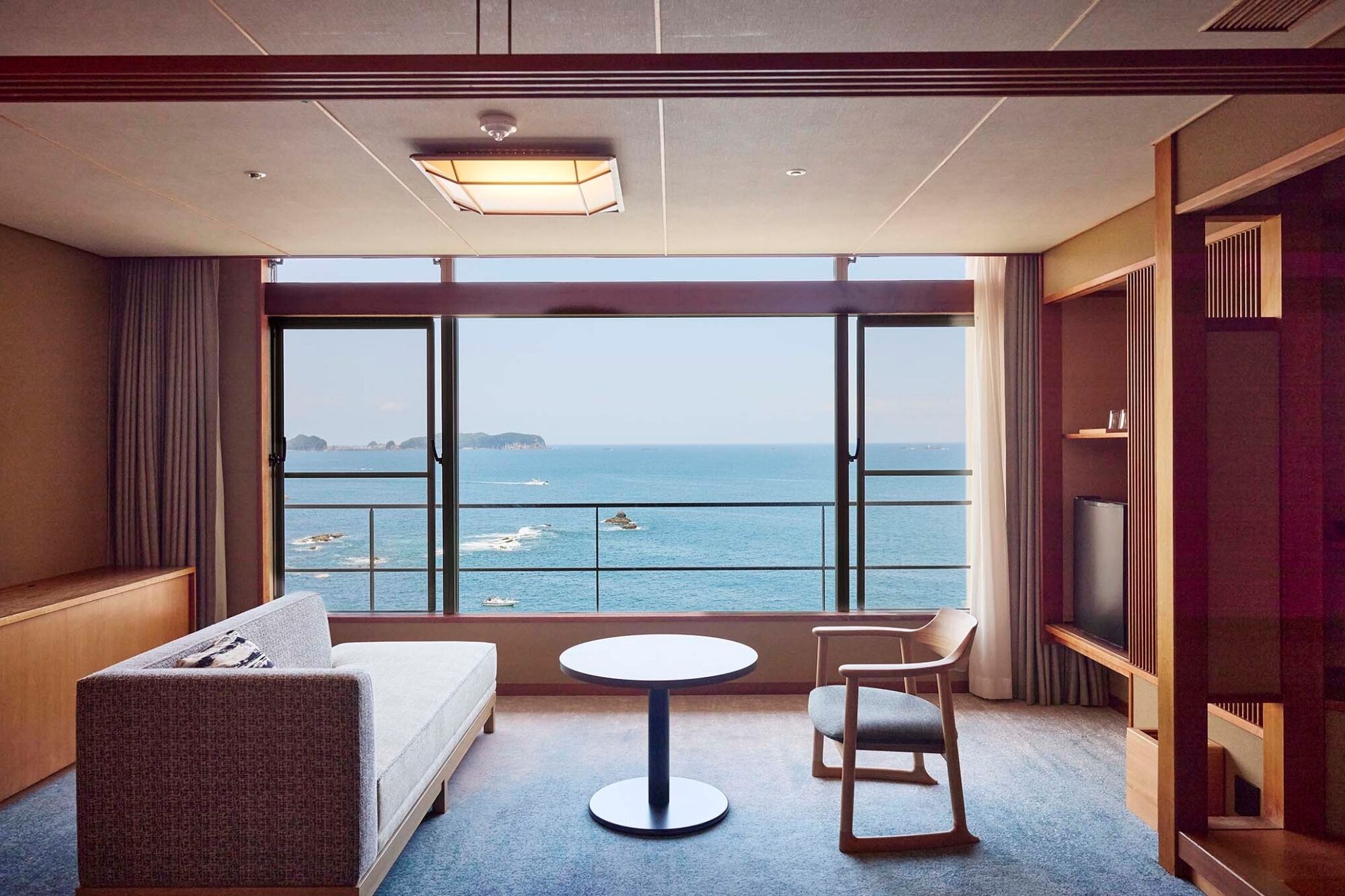 A hotel room with a large window offering a sea view, a sofa, a small round table, and a TV on a wooden stand.