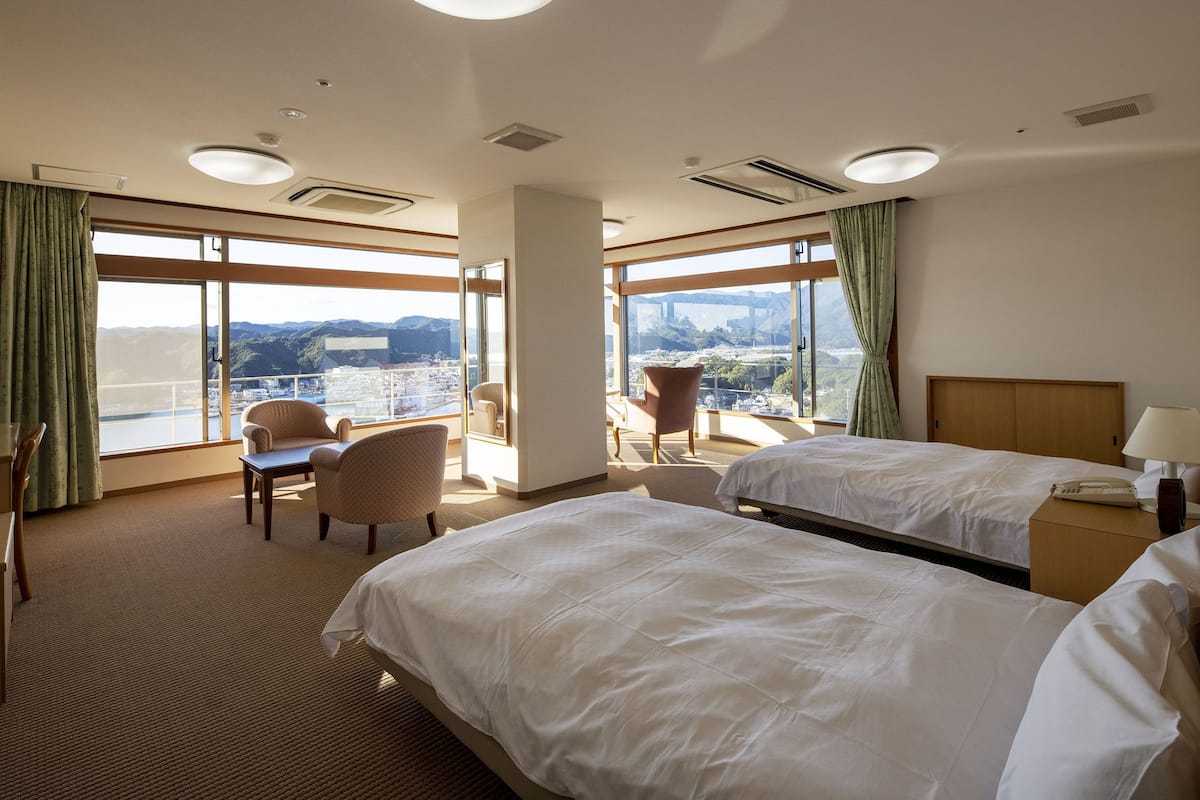 Une chambre d’hôtel avec deux lits, un bureau, une chaise et de grandes fenêtres donnant sur la ville.