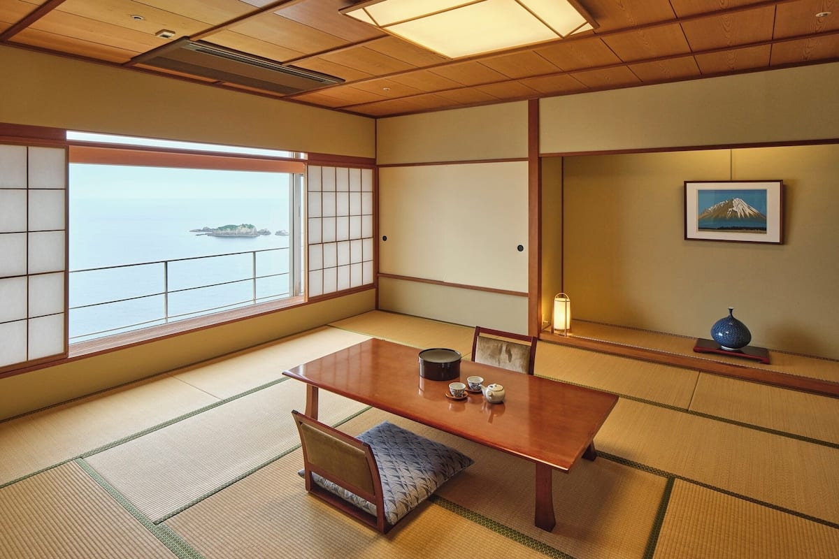 Une pièce de style japonais traditionnel, avec un sol recouvert de tatamis, une table en bois sur laquelle est disposé un service à thé, une vue sur la mer et un tableau représentant une montagne.