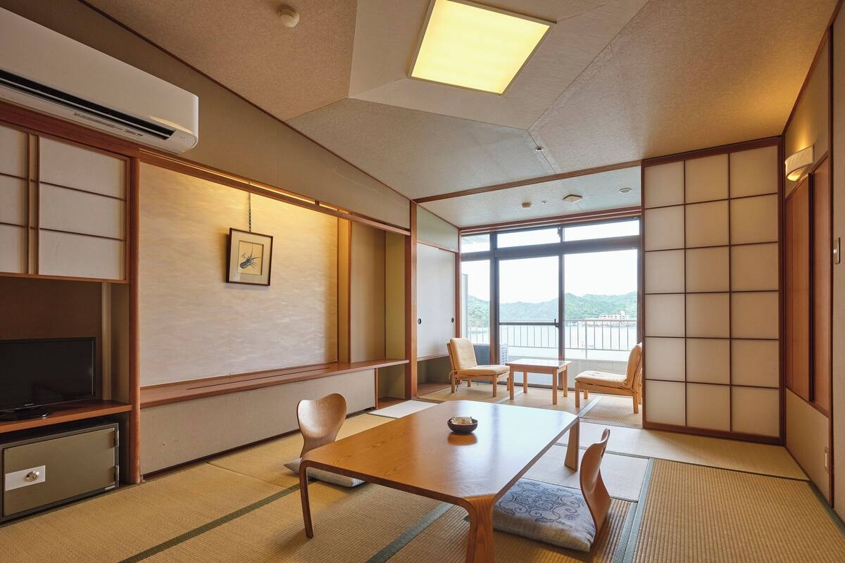 Une pièce de style japonais traditionnel, avec un sol recouvert de tatamis, une table basse, des chaises, un téléviseur et une vue sur une montagne, visible à travers de grandes fenêtres.