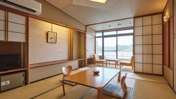 Een traditionele Japanse kamer met tatami vloerbedekking, een lage tafel, stoelen, een televisie en uitzicht op een berg door grote ramen.