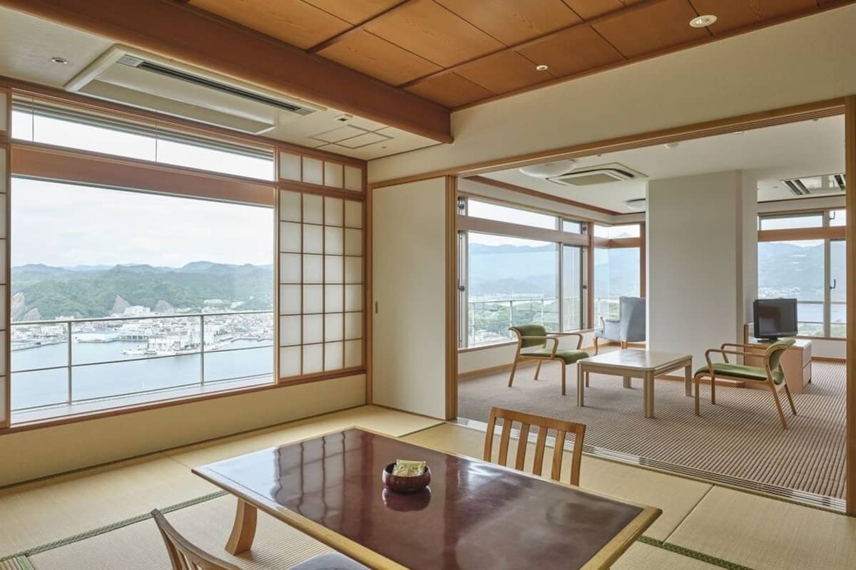 Une pièce de style japonais traditionnel, avec un sol recouvert de tatamis, une table basse et une vue sur le paysage urbain à travers de grandes fenêtres.