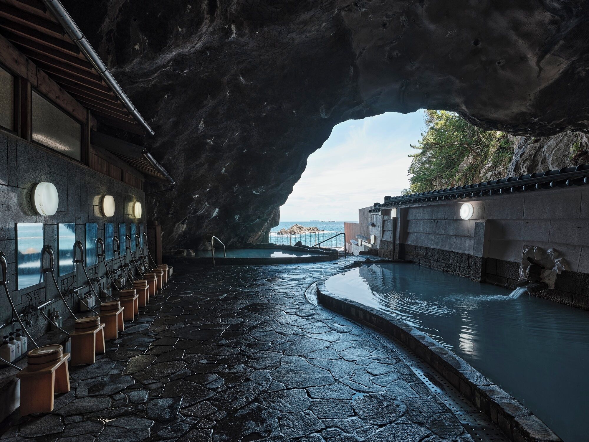 Uma área de spa com piso de pedra, piscina e vista para o mar através de uma estrutura semelhante a uma caverna.