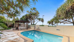 A swimming pool with lounge chairs, a covered seating area, and tropical plants.