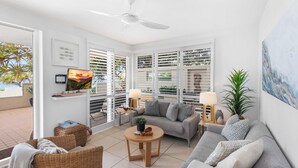 A bright living room with a grey sofa, wicker chairs, a round wooden table, and a ceiling fan.
