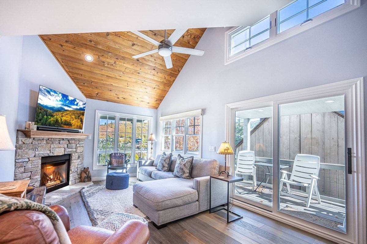 A living room with a stone fireplace, a ceiling fan, and a large sliding glass door leading to an outdoor area.