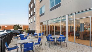Un espace extérieur aménagé en terrasse avec des tables et des chaises, une voiture garée à proximité et un bâtiment avec de grandes fenêtres en arrière-plan.