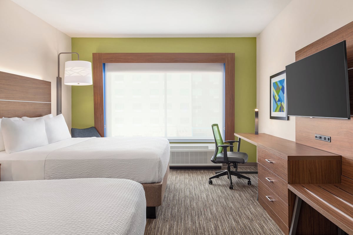 A hotel room with two beds, a desk, a chair, a TV, and a window with blinds.