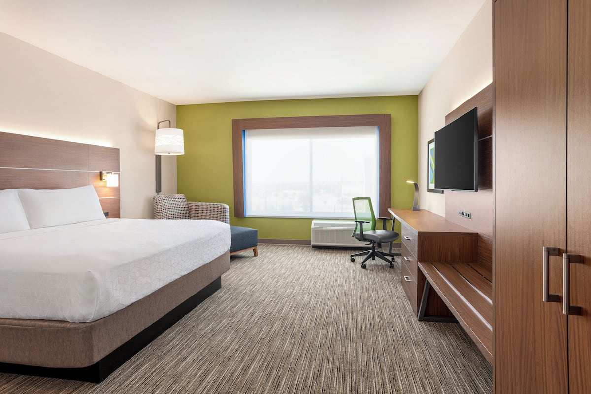 A hotel room with a large bed, a desk with a flat-screen TV, a chair, and a window with a city view.