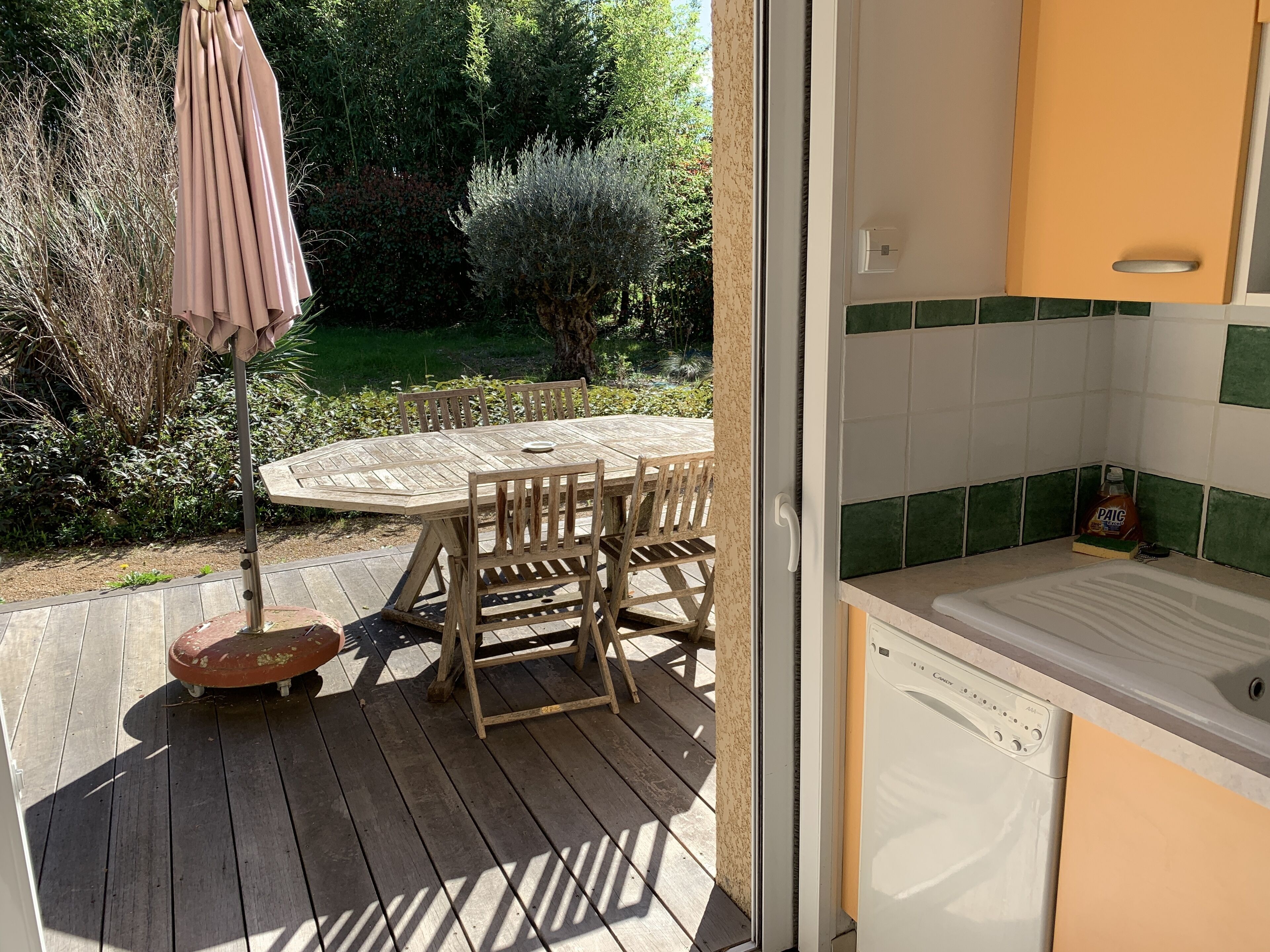 A wooden deck with a round table and chairs, a small stool, and a hanging umbrella. A kitchen area with a white dishwasher and green-tiled backsplash is visible through an open door.