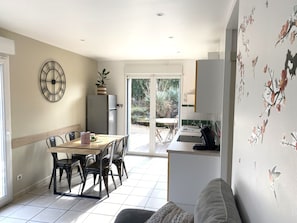 Un coin repas avec une table et des chaises, un réfrigérateur, une horloge et une vue sur une terrasse extérieure, accessible par une porte vitrée.