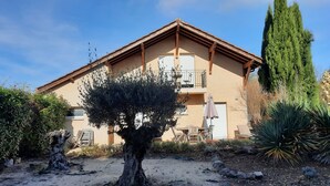 A two-story building with a balcony, a small garden with a tree, and a tall coniferous tree.