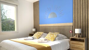 A neatly made bed with white bedding and a yellow throw, a nightstand with a lamp, and a wall with vertical wooden slats and a sun design.