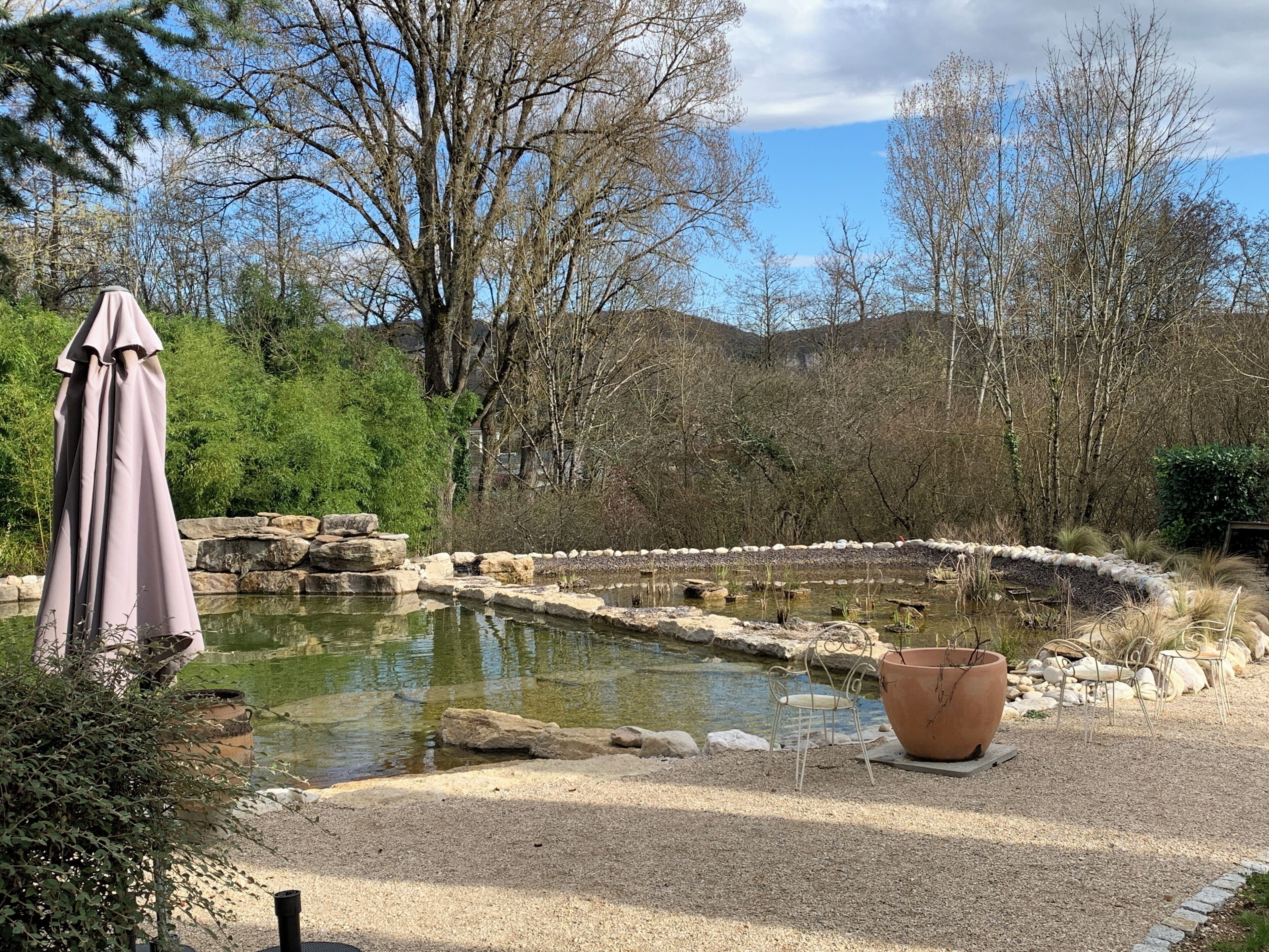 A tranquil outdoor setting with a pond, surrounded by rocks and greenery, and a seating area with a chair and a table.