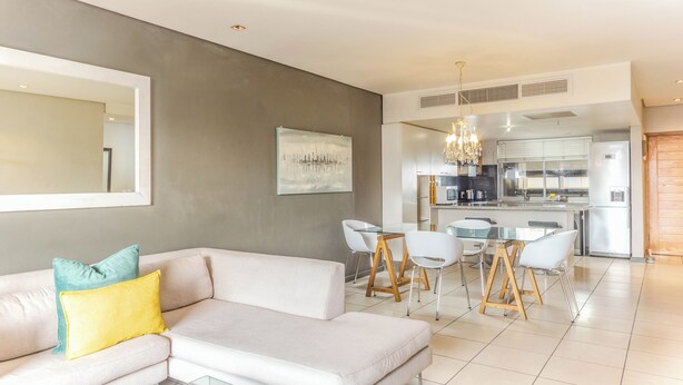 Una sala de estar moderna con un sofá, una mesa de comedor con sillas, una cocina al fondo y una araña.