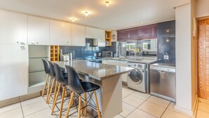 Una cocina moderna con una isla central, taburetes de bar, electrodomésticos integrados y un amplio ventanal con persianas.