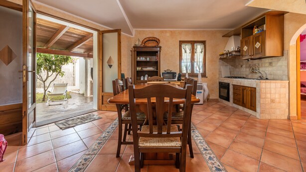 A kitchen with wooden cabinets, a dining table with chairs, and a view to an outdoor area. - Vacation Home Casa Giovanna e Antonio with Sea View, A/C, Terrace & Garden (Calasetta)