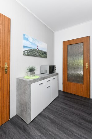 Un'accogliente area reception moderna, con una porta in legno, un mobiletto grigio con una pianta, un forno a microonde e un quadro con un paesaggio.