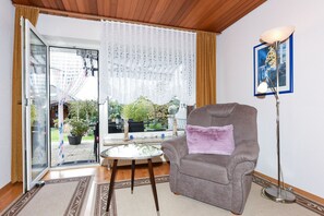Ein Wohnzimmer mit einem grauen Sessel, einem Glastisch, einer Stehlampe und Blick durch eine Schiebetür in einen Garten.