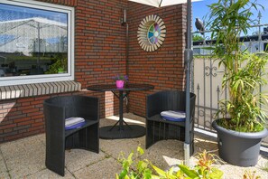 Eine Terrasse mit zwei Korbstühlen, einem runden Tisch, einem Blumentopf und einer Wanddekoration.