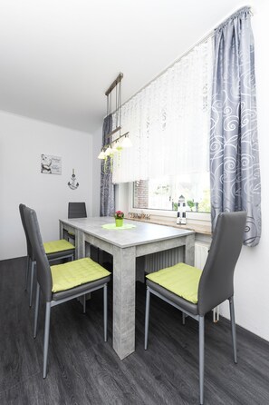 Et spiseområde med et gråt bord, fire stole med grønne hynder, et hængende lysarmatur og et vindue med mønstrede gardiner.