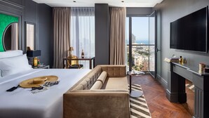 A hotel room with a bed, a sofa, a TV, a table with a bottle and glasses, and a view of the city. - Debrah Brown (Tel Aviv)