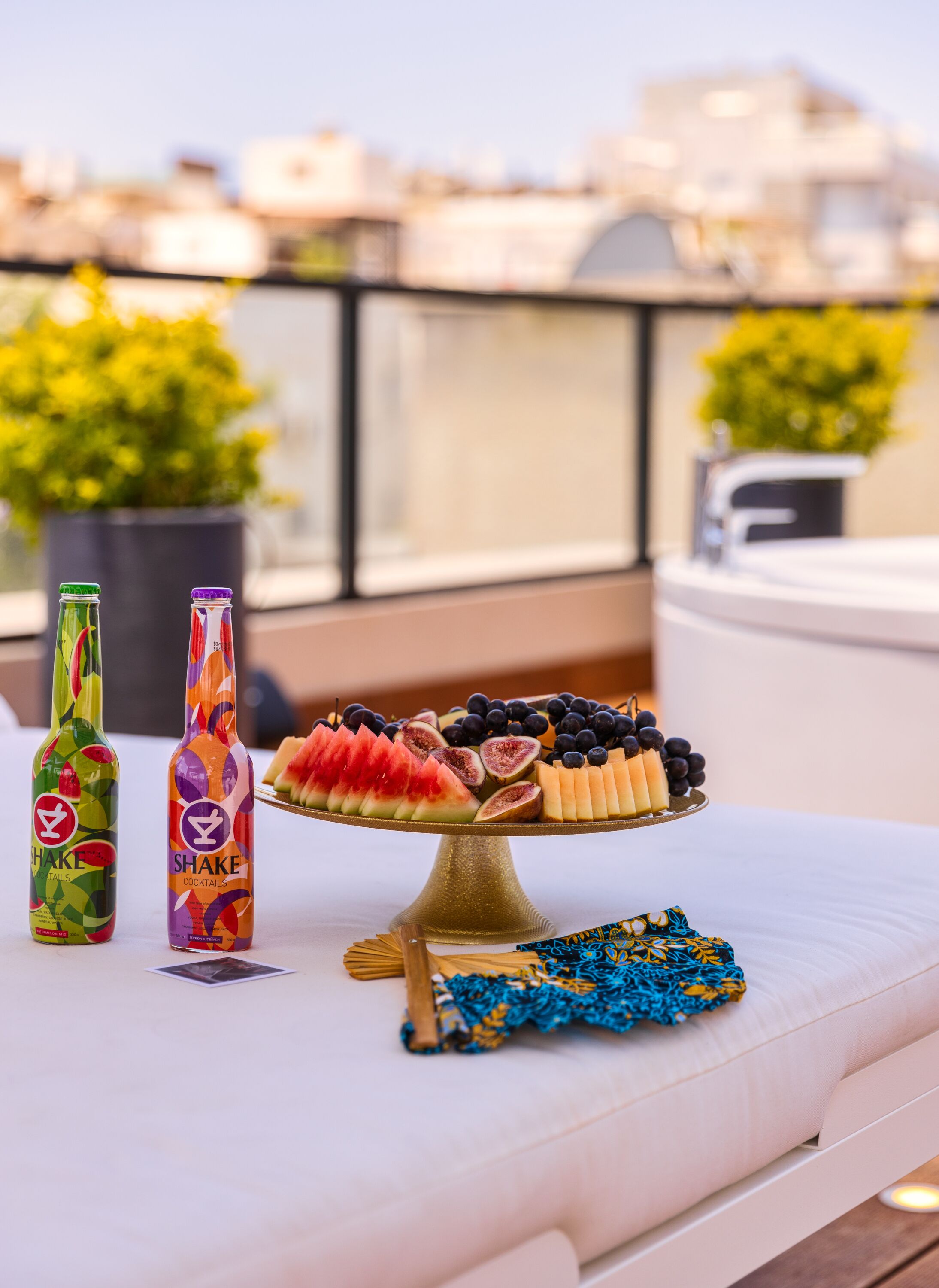 A table with two bottles labeled 'SHAKE' and a fruit platter on a balcony with a city view.