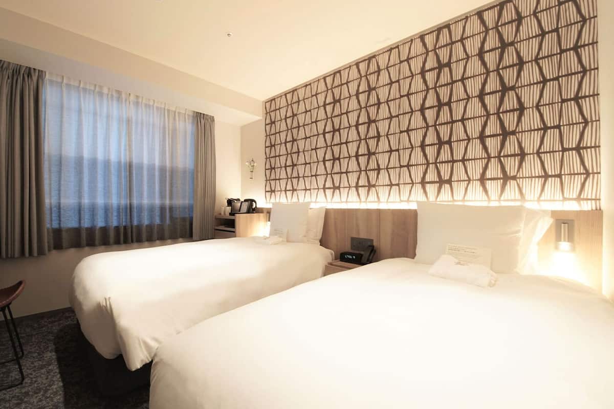 Une chambre d’hôtel avec deux lits, un bureau, une chaise et une grande fenêtre avec des rideaux.