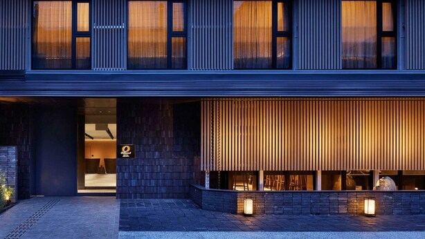 A modern hotel entrance with a dark facade, large windows, and a visible reception area.