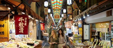 A narrow market street with stalls selling various goods, illuminated by hanging lanterns and signs.