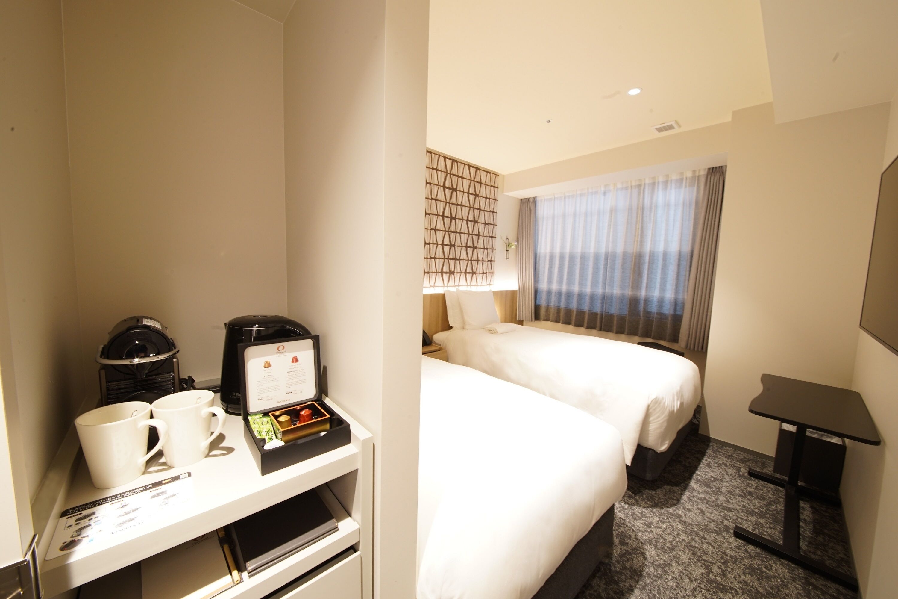 Ein Hotelzimmer mit zwei Betten, einem kleinen Schreibtisch mit Kaffeemaschine und Tassen sowie einem Fenster mit Vorhängen.