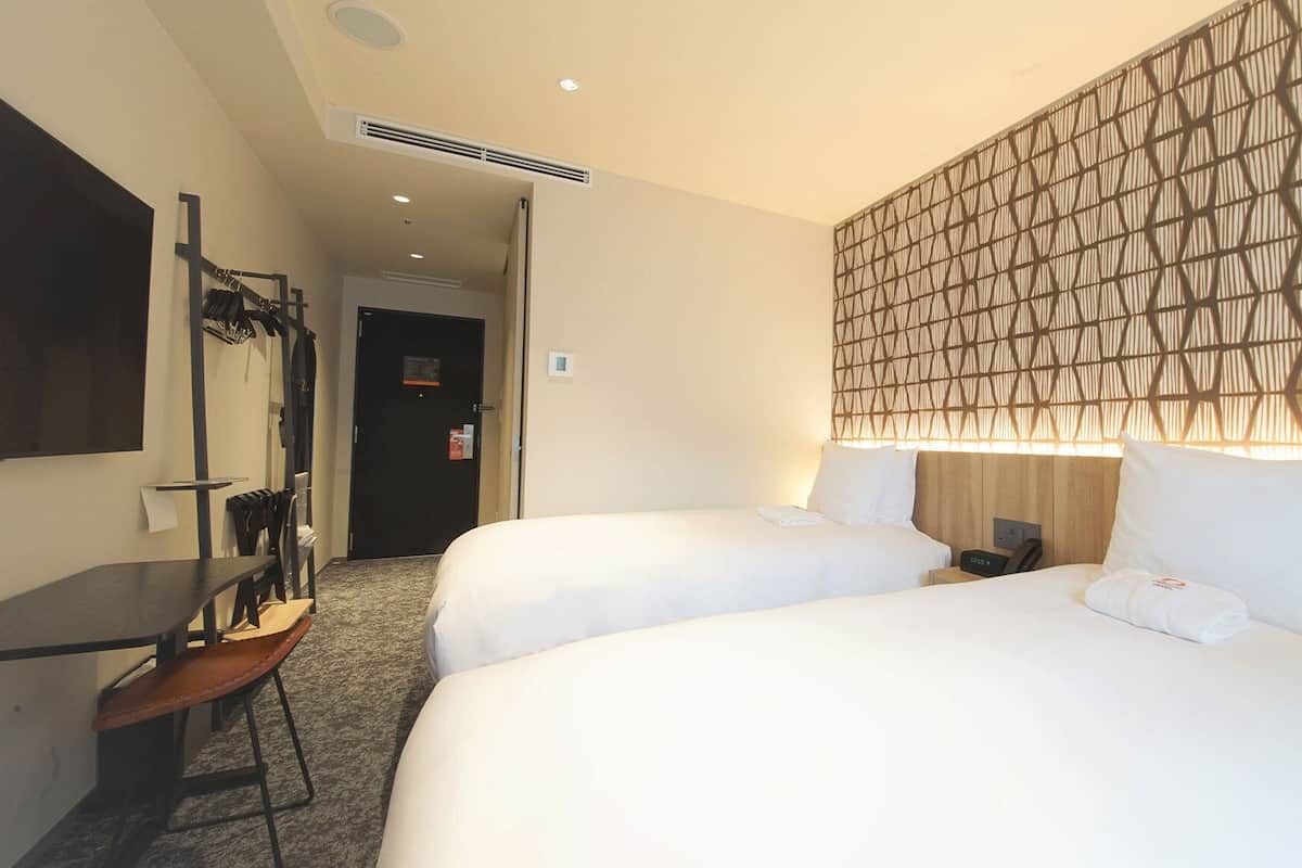 Une chambre d’hôtel avec deux lits, un bureau et une chaise. Il y a un téléviseur fixé au mur et une porte qui donne sur une autre pièce.