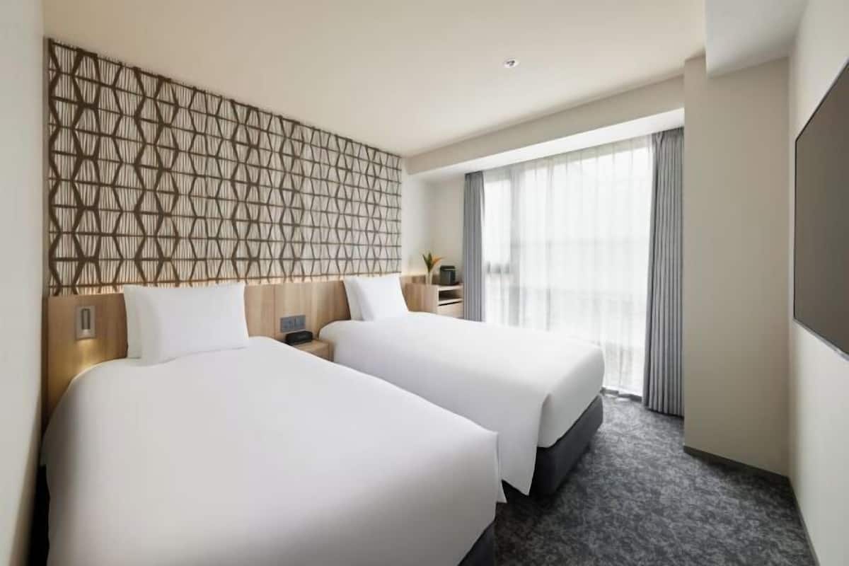 Une chambre d’hôtel avec deux lits, une grande fenêtre avec des rideaux, un téléviseur fixé au mur et un mur orné d’un motif géométrique.