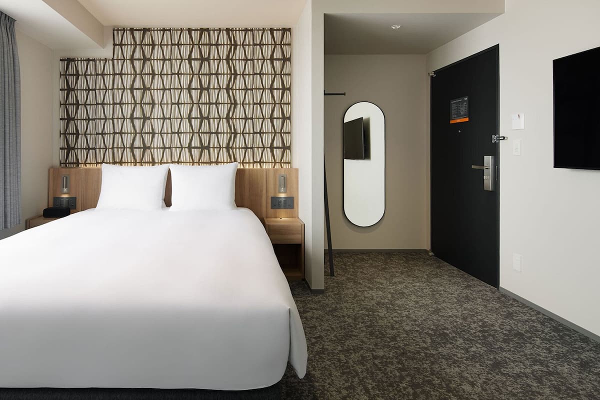 Une chambre d’hôtel moderne avec un grand lit, une tête de lit en bois, un miroir fixé au mur, une porte et un sol recouvert de moquette.