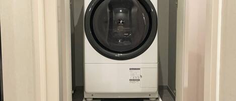 A compact, white front-loading washing machine in a narrow hallway with tiled flooring.