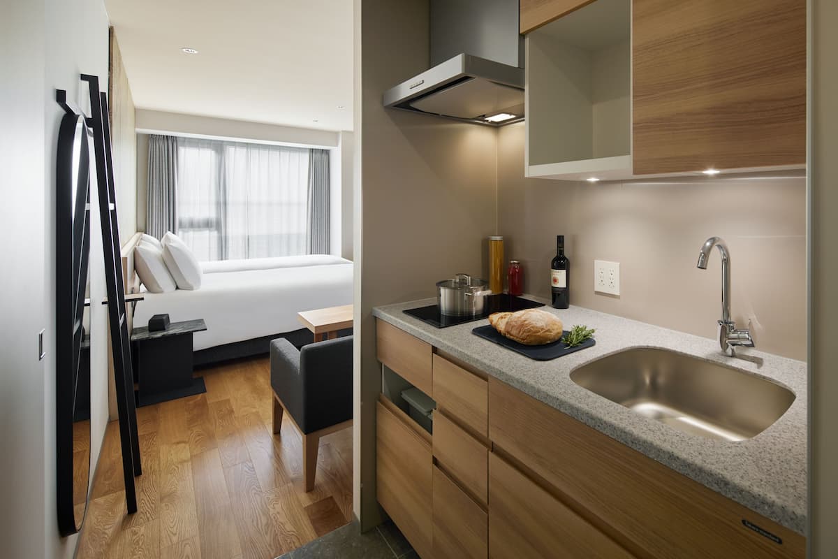 Une chambre d’hôtel de taille réduite, comprenant un lit, une petite kitchenette, un évier et une fenêtre avec des rideaux.