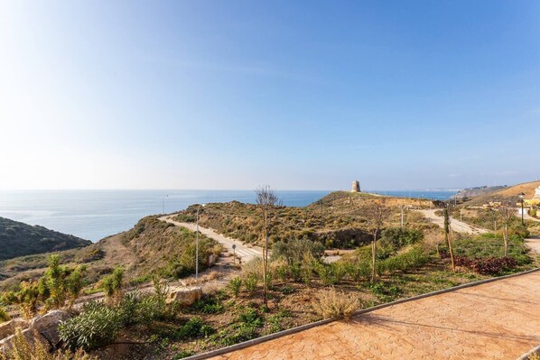 Un paisaje costero con un camino pavimentado, vegetación escasa y una torre distante.