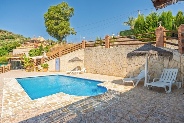 A swimming pool with a stone-tiled deck, white lounge chairs, and a stone wall with a thatched umbrella. - Villa Olympo by Solset (Torrox)