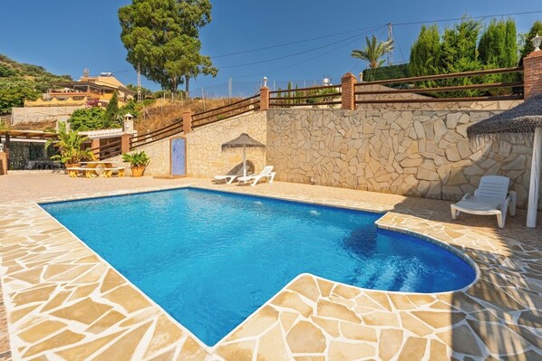 A swimming pool with stone tiles, white lounge chairs, and a stone wall with a wooden fence. - Villa Olympo by Solset (Torrox)