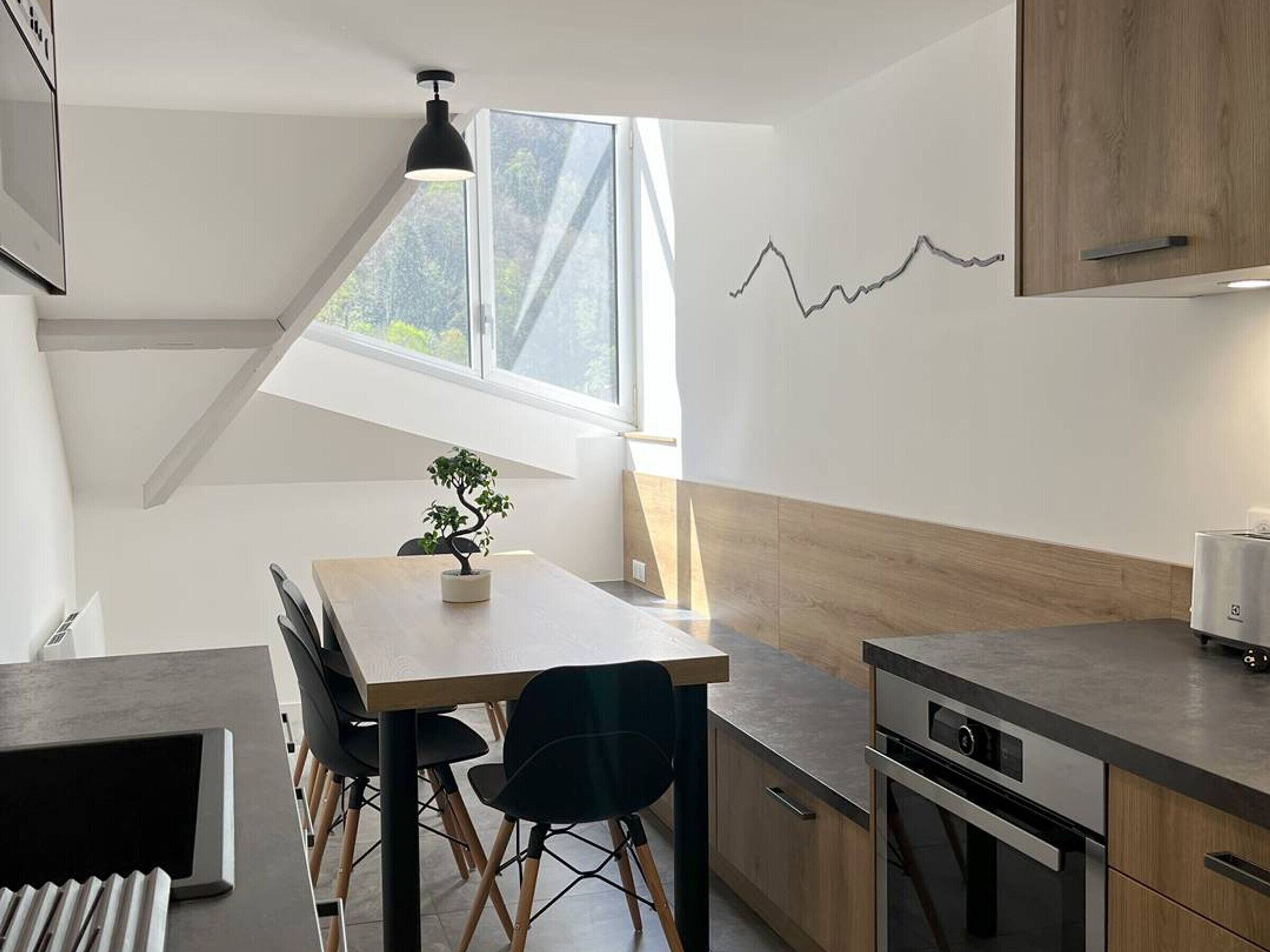 Een moderne keuken met een eethoek, met een houten tafel, stoelen en een kleine potplant. Een raam zorgt voor natuurlijk licht, en er is een wandlamp te zien.