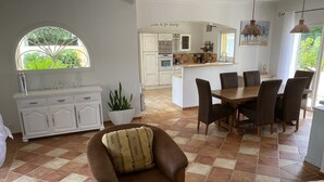 Zona d'estar àmplia amb una butaca de cuir marró, una taula de menjador amb cadires, una cuina al fons i una finestra gran amb vistes a la vegetació.