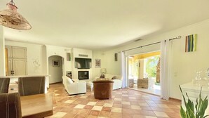 Una zona d'estar espaiosa amb un paviment a quadres, un sofà blanc, un televisor muntat sobre una llar de foc, i una zona de menjador amb taula i cadires.