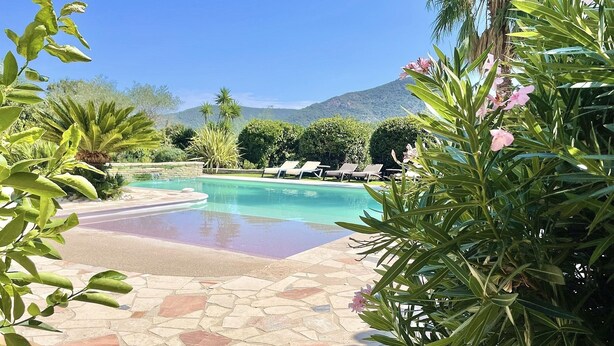 Zona de piscina amb terra de rajola, envoltada de vegetació i una muntanya al fons.
