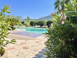 A pool area with a tiled floor, surrounded by greenery and a mountain in the background.